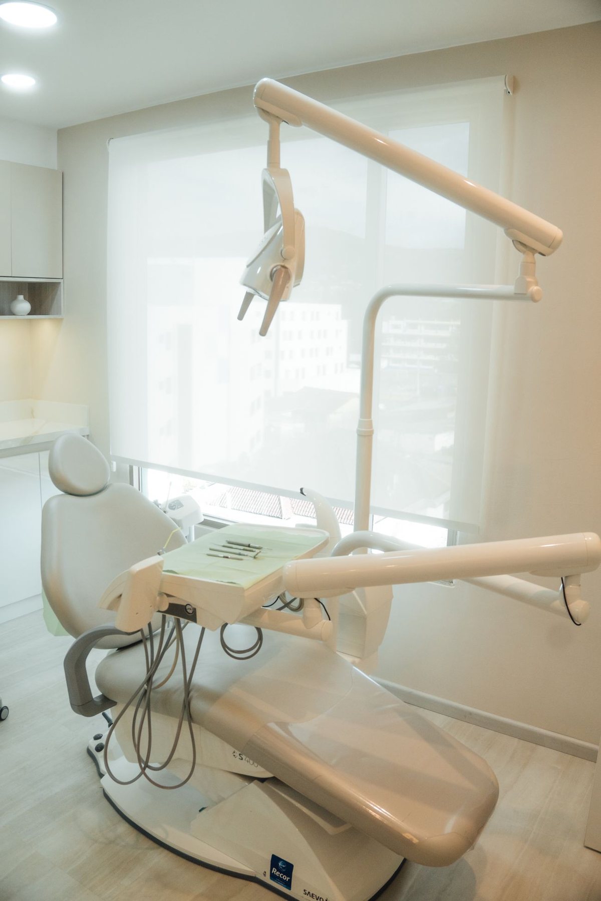 A quiet dental treatment room in soft daylight, with a cream chair beside a shaded window.