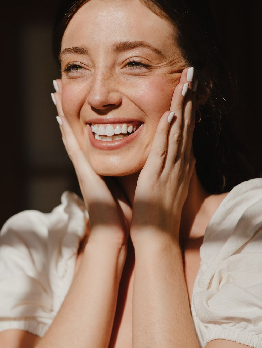 A person smiling easily in warm daylight, hands resting against their cheeks.